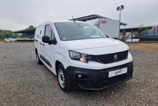 Peugeot Partner blanc vue 3/4 avant droit sur parking, avec calandre noire et feux allumés.