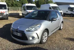Citroën DS3 gris clair métal vue 3/4 avant droit sur un parking avec plusieurs fourgons blancs en arrière-plan.