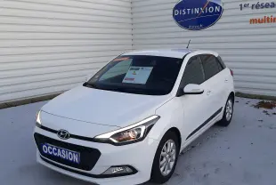 Vue 3/4 avant droite d'une Hyundai i20 blanche de 2016 avec phares allumés, stationnée devant un mur blanc.
