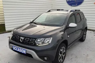 Dacia Duster gris Cassiopée en 3/4 avant droit, avec jantes bi-ton et barres de toit visibles.