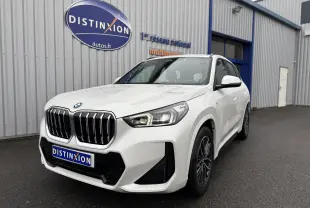 BMW X1 blanc minéral en 3/4 avant droit, avec calandre noire et phares LED distinctifs, stationné devant un bâtiment.