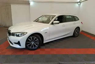 BMW Série 3 320e blanche vue 3/4 avant droit en break, avec jantes alliage et prise de recharge latérale visible.