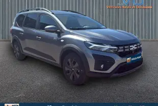 Vue 3/4 avant d'un Dacia Jogger gris 2025 avec barres de toit et jantes spécifiques, stationné sur sol goudronné.
