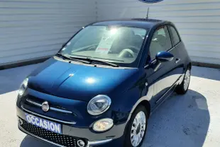 Vue 3/4 avant d'une FIAT 500 bleu foncé avec phares ronds et jantes blanches, stationnée devant un mur blanc.