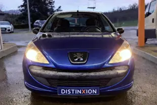 Peugeot 206+ bleu vue 3/4 avant droit avec phares allumés, garée devant un bâtiment vitré.