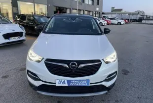 Vue avant d’un Opel Grandland X blanc avec toit noir, phares allumés, sur parking extérieur devant un bâtiment.