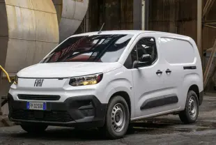 Fiat Doblo fourgon blanc vu en 3/4 avant droit dans un environnement industriel, avec calandre et phares allumés.
