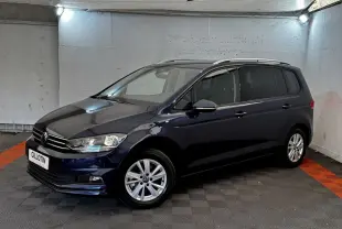 Volkswagen Touran bleu 3/4 avant droit en intérieur, avec jantes alliage et rails de toit chromés visibles.