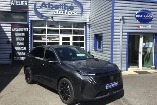 Peugeot 3008 gris titane en 3/4 avant droit, avec calandre noire et jantes alliage noires devant un garage automobile.