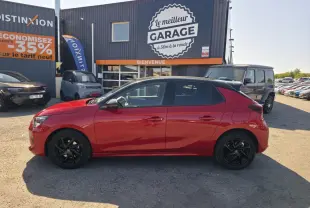 Opel Corsa rouge vue de profil côté gauche, avec toit noir et jantes alliage noires, stationnée devant un garage.