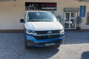 Vue avant d'un Volkswagen Transporter blanc avec pare-chocs bleu, stationné devant un bâtiment commercial.