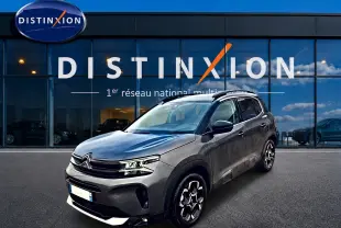 SUV Citroën C5 Aircross gris vu en 3/4 avant droit, avec jantes alu et barres de toit visibles devant un showroom.