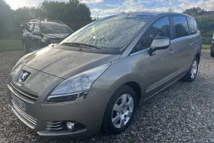 Peugeot 5008 2013 gris métallisé vue 3/4 avant droit sur un sol gravillonné avec ciel nuageux.