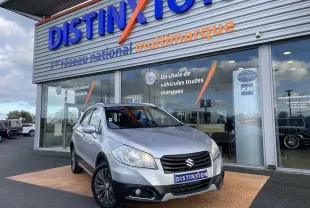 Suzuki SX4 S-Cross gris métallisé, vue 3/4 avant droit, avec projecteurs allumés et calandre noire distinctive.