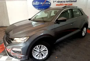 Volkswagen T-Roc gris foncé vue 3/4 avant droit, avec jantes argentées et phares allumés en intérieur showroom.