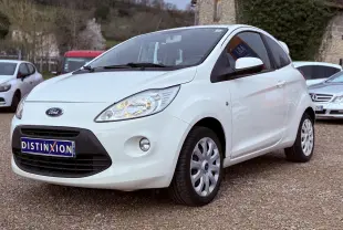 Ford Ka blanc vue 3/4 avant droit sur parking, avec phares allumés et jantes tôles blanches.