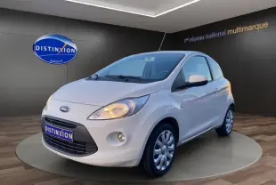 Vue 3/4 avant droite d'une Ford KA blanche de 2015 avec phares allumés, sur fond intérieur showroom.