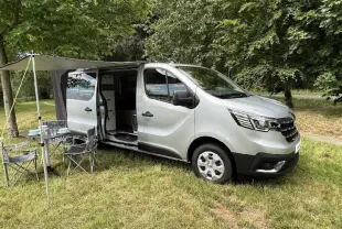 Renault Trafic Fourgon gris en 3/4 avant droit avec porte latérale ouverte et auvent déployé en pleine nature.
