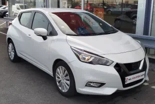 Nissan Micra blanche vue 3/4 avant droit, avec phares allumés et jantes alliage, stationnée devant un showroom.