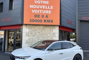 DS4 blanc vue 3/4 avant droit, jantes noires, calandre distinctive, devant un bâtiment avec panneau orange Distinxion.