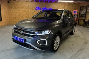 Volkswagen T-Roc gris 3/4 avant droit, phares allumés, calandre chromée et jantes alliage visibles en intérieur showroom.