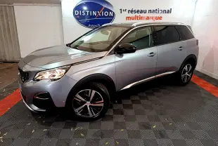 Peugeot 5008 gris clair vue 3/4 avant droit, avec jantes alliage et barres de toit chromées en intérieur showroom.