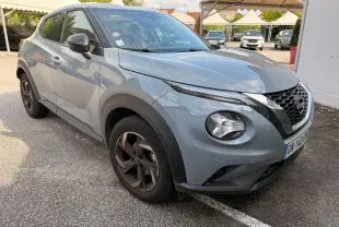 Nissan Juke gris clair vu en 3/4 avant droit, avec jantes noires et calandre distinctive en V chromé.