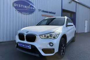 BMW X1 blanc vue 3/4 avant droit avec phares LED allumés devant un bâtiment industriel.