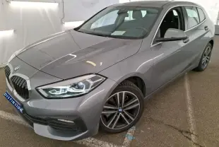 BMW Série 1 gris vue 3/4 avant droit, avec jantes alliage et phares LED, en intérieur showroom.