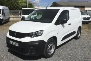 Vue 3/4 avant d’un utilitaire Peugeot Partner blanc avec pare-chocs noirs et jantes acier, stationné sur un sol gravillonné.