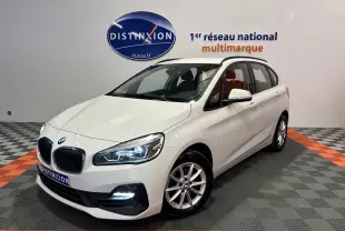 BMW Série 2 Active Tourer 216d blanc vue 3/4 avant droit avec phares allumés dans showroom.