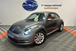 Volkswagen Coccinelle gris vue 3/4 avant droit avec phares allumés dans showroom lumineux.