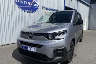 Vue 3/4 avant d'un Citroën Berlingo gris Artense métal avec calandre noire et jantes foncées, stationné devant un bâtiment.