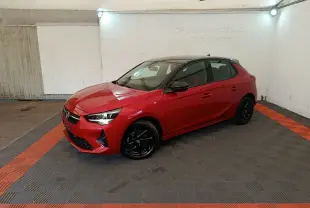 Opel Corsa rouge 2023 vue 3/4 avant droit, toit noir, jantes alliage noires, dans un garage éclairé.
