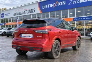 Vue 3/4 arrière d’un Nissan Qashqai 2024 rouge foncé avec toit noir et jantes noires, stationné devant un garage.