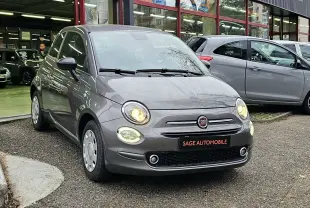 Fiat 500 gris vue 3/4 avant droit, phares allumés, stationnée devant un garage Sage Automobile.