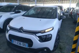 Vue 3/4 avant d'une Citroën C3 Société blanche de 2021 avec feux allumés, garée sous abri sur sol gravillonné.