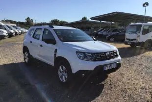 Dacia Duster blanc vu en 3/4 avant droit sur un terrain avec plusieurs véhicules en arrière-plan.
