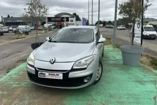 Renault Mégane 3 gris vue de face, légèrement décalée côté gauche, sur parking extérieur avec panneaux publicitaires.