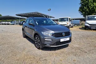 Volkswagen T-Roc gris foncé métal en 3/4 avant droit sur parking extérieur avec plusieurs véhicules utilitaires en arrière-plan.