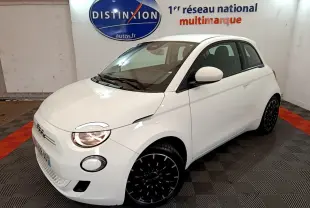 Fiat 500 électrique blanc vue 3/4 avant droit en intérieur avec jantes noires et phares allumés.