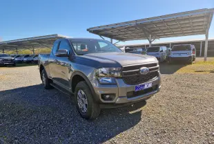 Vue 3/4 avant droit d’un Ford Ranger Super Cabine gris carbone diesel, stationné sur un terrain gravillonné en plein jour.