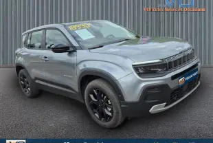 Jeep Avenger gris foncé en 3/4 avant droit, avec jantes noires et calandre distinctive, stationné devant un mur métallique.