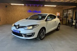 Renault Mégane Coupé blanc vue 3/4 avant droit en intérieur avec phares allumés et jantes alliage.