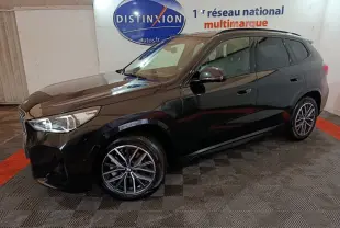 BMW X1 xDrive23i M Sport noir vue 3/4 avant droit dans un garage avec sol à damier et logo Distinxion en arrière-plan.
