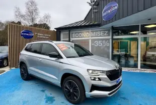 Skoda Karoq gris diamant métallisé en 3/4 avant droit, avec toit panoramique et jantes noires, devant un concessionnaire.