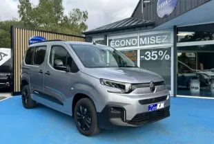 Vue 3/4 avant gauche d'un Citroën Berlingo gris acier avec jantes noires, stationné devant un magasin.