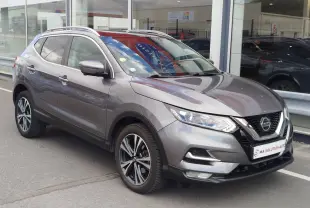 Nissan Qashqai gris Squale en 3/4 avant droit, avec jantes alliage et calandre chromée visible devant un showroom.