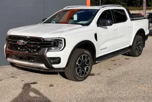Ford Ranger Wildtrak blanc vu en 3/4 avant droit, avec jantes alu noires et barres de toit visibles.