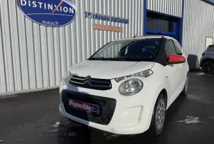 Citroën C1 blanc vue 3/4 avant droit avec rétroviseurs rouges, stationnée devant un bâtiment commercial.